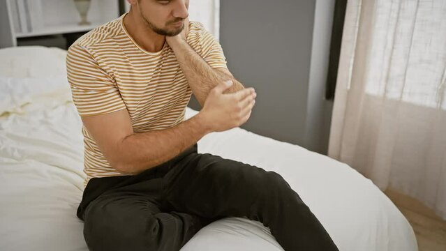 Hispanic man applying lotion to elbow in a cozy bedroom setting, conveying a personal care routine.