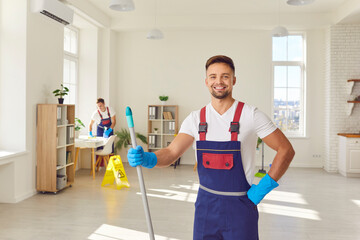 Portrait of happy smiling man janitor from professional cleaning service in uniform looking cheerful at camera holding mop in hand mopping house or office. Housework and housekeeping concept.