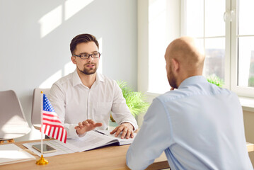 Serious male Consul of United States of America sitting at office table with American flag and...