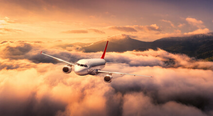 Plane is flying over mountain peaks in low clouds at golden sunrise. Top view of passenger...