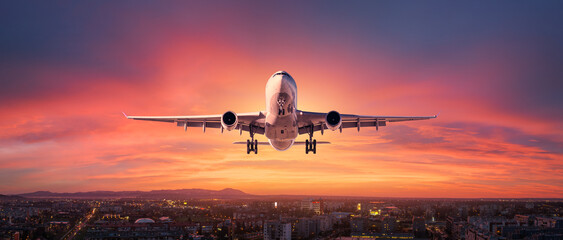 Airplane is flying in colorful sky over city at sunset. Landscape with passenger airplane, skyline, purple sky with red and pink clouds at dusk. Aircraft is landing at twilight. Aerial view of plane - obrazy, fototapety, plakaty