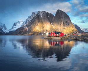 Red rorbuer on the island against snowy rocks lighted by sun, blue sea and dramatic cloudy sky in winter. Landscape with houses, mountains at sunset. Rorbu in Hamnoy village, Lofoten islands, Norway - 758213671