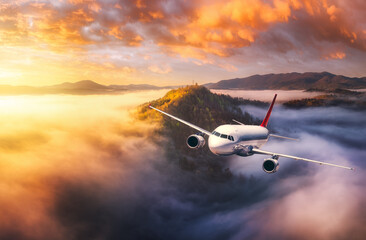 Plane is flying over mountain peaks in low clouds at golden sunrise. Top view of passenger...