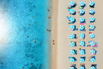 Aerial view of beautiful umbrellas on white sandy beach, people in blue sea on summer sunny day. Beach, Sardinia, Italy. Tropical landscape with azure water. Travel and vacation. Top view of seaside