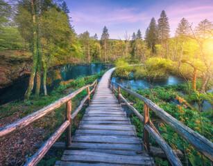 Wooden path in green forest in Plitvice Lakes, Croatia at sunset in spring. Colorful landscape with stairs in blooming park, trees, water lilies, river, pink sky in summer. Trail in woods. Nature - 758232648