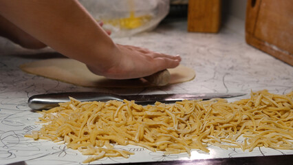 Hands are rolling out egg dough on a patterned work surface, with a tray of freshly cut light yellow pasta in the background.