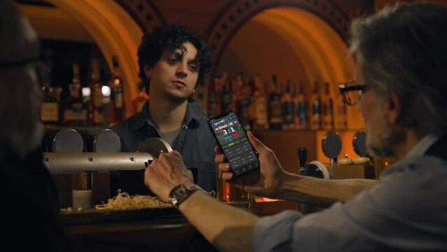 Close up of sports fan watching game score and bookmaker ratings using mobile phone. Man bets on football world league match online in app sitting at bar counter in pub at night. Gambling concept.