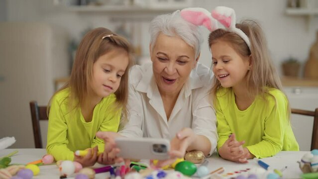 Easter grandmothers with granddaughters. Smiling grandmother with twins grandchildren watching cartoons on phone in rabbit bunny ears, talking, celebrating at home. Easter family holiday concept.