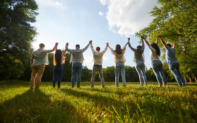 Group of friends having fun in nature. Back view team of several young diverse people standing in a row on a green grassy lawn in the park, holding hands and raising them up all together