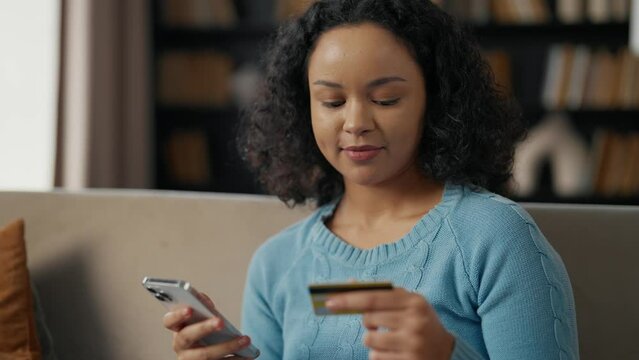 African american woman doing online payment. Young girl buys in internet on smartphone using credit card, enters card number in marketplace sitting on sofa at home. Online purchases, customer concept.