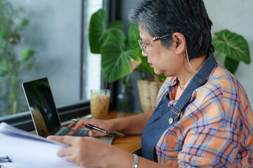 Gray-haired woman, pensioner, businessman, owner of small cafe Sitting in front of notebook holding pen to check store's sales documents,  serious expression on face. Next to it, coffee behind it.