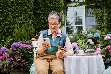 mature cheerful woman sitting in garden at tea time and looking at phone near house in England