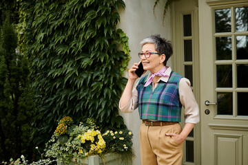 mature cheerful elegant woman with glasses and short hair talking by her phone near house in England