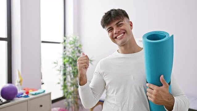 Handsome young hispanic man flashes a cheerful smile, pointing sideways with a thumb-up gesture, as he holds his yoga mat, ready for a friendly rehab clinic session. joyful indoor expression.