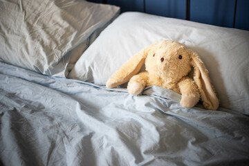 White bunny plush toy laying under covers in parents bed with blue blankets and soft day light. Side view, background with copy space.