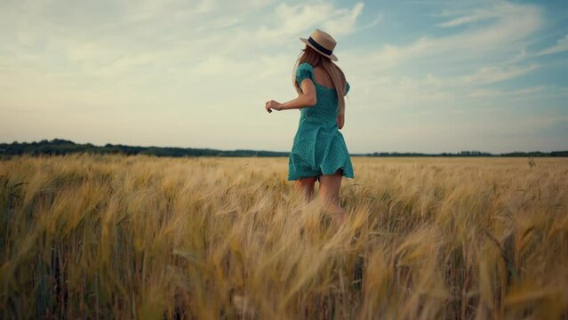 Carefree woman in straw hat, blue dress running on wheat field feeling freedom, harmony, lightness and serenity, back view. Smiling blonde female enjoying nature resting on farm, nature activity.