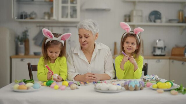 Easter grandmothers with granddaughters. Smiling grandmother with twins grandchildren painting decorating eggs in rabbit bunny ears, celebrate together at home. Easter decorations together concept.