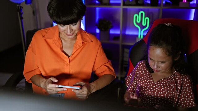 Mother-daughter streamer duo lighting up the gaming room, playing video game on smartphone embellishing family love