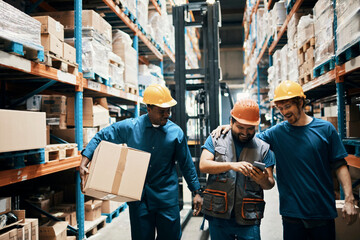 Happy warehouse workers looking at phone, men with hard hats in storage facility