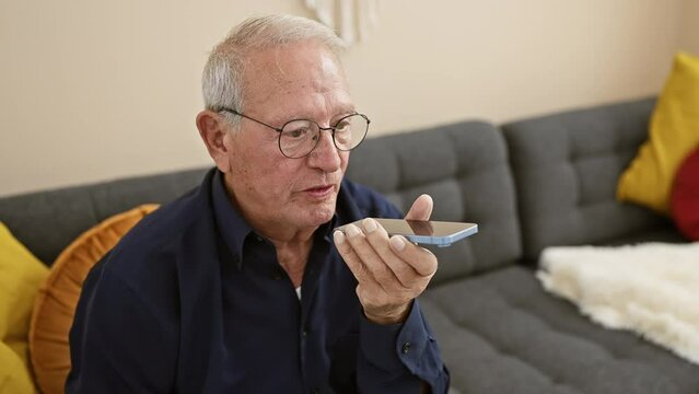 Confident senior man smiling happily while sending audio message using smartphone, sitting on home sofa