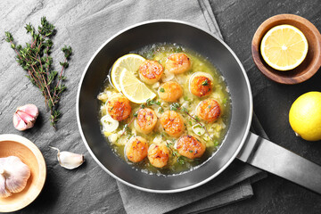 Delicious scallops with sauce in frying pan and ingredients on dark gray textured table, flat lay