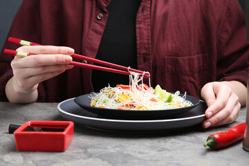 Stir-fry. Woman with chopsticks eating tasty rice noodles with meat and vegetables at grey textured table, closeup