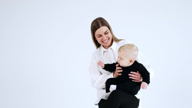 Happy smiling woman in white shirt holding a blond baby wearing black suit. Child with cute plump cheeks sits peacefully on mom's laps.