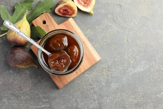 Jar of tasty sweet jam, fresh figs and green leaf on grey table, flat lay. Space for text