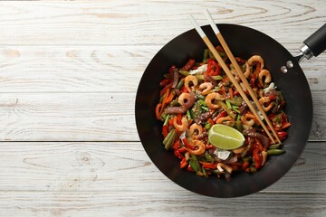 Shrimp stir fry with vegetables in wok and chopsticks on light wooden table, top view. Space for text