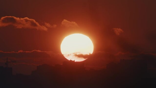 A breathtaking sunset, with the sun's orb partially obscured by clouds, descends over a darkened cityscape.