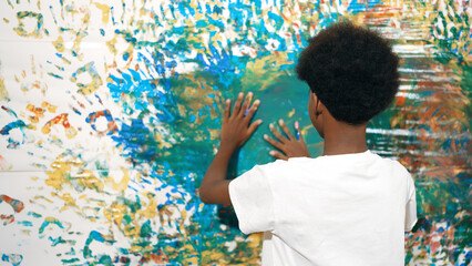 Back view of happy african boy painted the stained wall with colorful hand while wearing casual white shirt in art lesson.Smart student use hand print to make creative artwork. Education. Edification. © InfiniteFlow