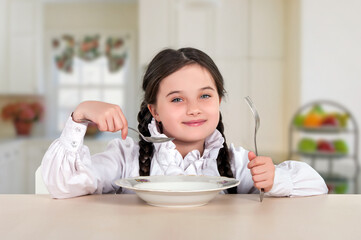 little teenage girl sitting at the dining table in the kitchen. Before appetite eating with a plate, spoon. 