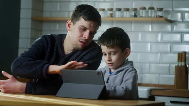 Young dad and son having fun using digital tablet sitting in the kitchen. Cute kid learning technology look at pad screen at home.
