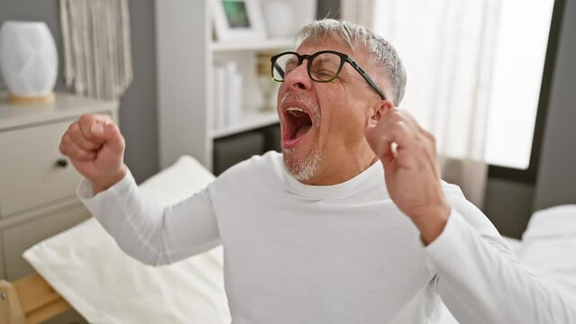 Tired, yawning middle-aged grey-haired man in pyjamas, overcome with sleepiness, covers mouth with hand in a bedroom -- a portrait of boredom and fatigue.