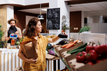 Woman in zero waste shop purchasing organic locally grown vegetables, picking ripe eggplants. Customer in local grocery shop looking to buy healthy food, using paper bag to avoid single use plastics
