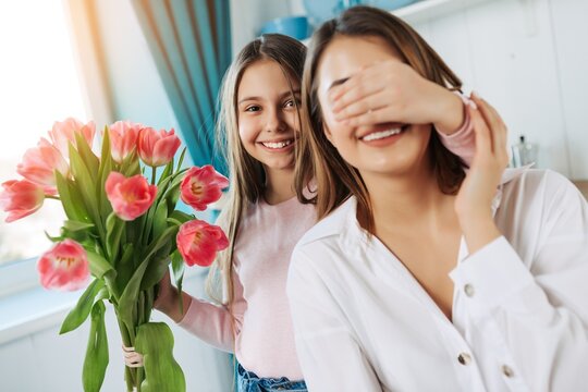 Happy Mother's Day! Cute charming girl closing mother`s eyes to make a surprise and give a bouquet of tulip flowers on kitchen