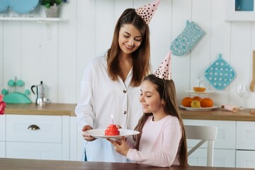 Cute little girl blowing out candles on birthday cake on kitchen with mother. Celebrating birthday at home