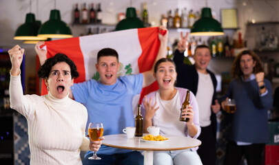 Company of emotional adult young sports fans holding flag of Peru, rejoicing winning match game and...