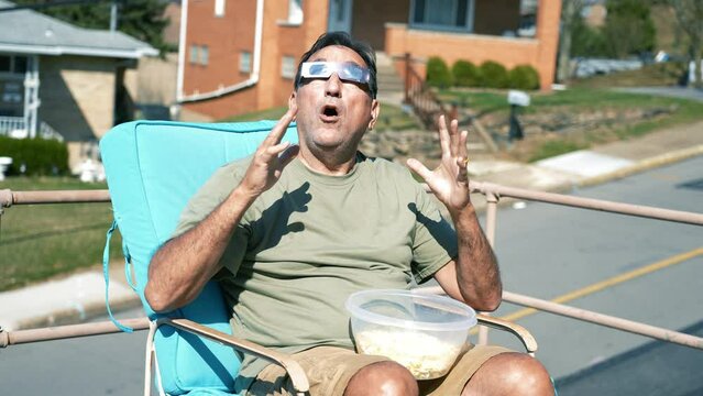 A wide shot of a man eating popcorn while looking at a total solar eclipse while wearing protective eclipse sunglasses.  	
