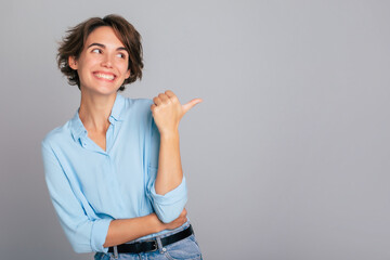 Shot of excited happy female in blue shirt demonstrating and pointing on copy space adverts isolated over gray background - 758377640