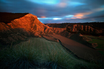 The last rays of sunlight cast a fiery glow on the textured hills of Central Guadalajara, with long grasses in the foreground and a dynamic sky above