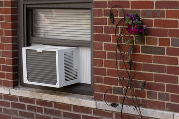 exterior view of air conditioning window unit extruding from the window sill of a red brick building with a deorative flower next to it