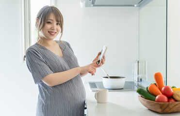 キッチンでスマートフォンを使う妊婦の女性（マタニティ・アプリ・栄養管理・食事管理・食事記録）
