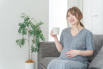 家のソファでホットドリンクを飲む妊婦の女性・マタニティ・プレママ
