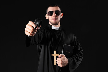 Young priest with gun and Holy Bible on black background