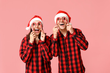 Funny young couple in Christmas pajamas and with candy canes on pink background