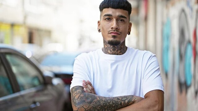 Cool young latin man with tattooed arms, standing outdoors on the urban street, carrying a relaxed yet serious expression with his arms casually crossed under the sunny city skyline.