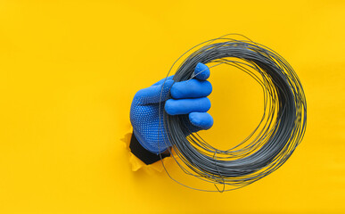 A left man's hand in a blue knitted glove holds a coil of steel wire. Torn hole in yellow paper. The concept of a worker, a labor migrant, a master of his craft. Copy space. - Powered by Adobe