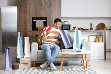 Young man with gift voucher and shopping bags sitting on sofa at home