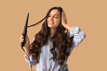 Beautiful young woman with curling iron on beige background